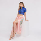 May include: A royal blue short-sleeved crop top with a front zipper and a small white flower graphic, paired with a light pink sarong-style skirt featuring a green leaf and orange flower print, finished with a delicate fringe along the bottom hem.