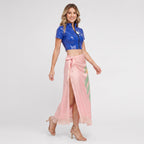 May include: A blue short-sleeved cropped top with a front zipper and a white floral graphic, paired with a light pink wrap-style maxi skirt featuring a high side slit, a tie waist, a green leaf pattern, and a fringed hemline with beige sandals.
