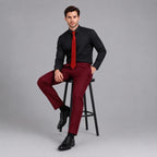 May include: A long-sleeved black button-down shirt paired with a bright red necktie and deep burgundy trousers. The outfit is completed with polished black leather dress shoes, creating a formal ensemble that is worn while seated on a black stool.