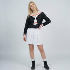 May include: A black long-sleeve top with a white sailor-style collar featuring orange stripes, paired with a white pleated mini skirt. The top includes a matching orange emblem on the chest and sleeve, and the outfit is worn with black boots.