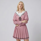 May include: A dusty pink long-sleeved dress featuring a white sailor-style collar with black trim, a black ribbon tie at the neckline, and a pleated skirt. The bodice includes double-breasted buttons and the sleeves have buttoned cuffs.